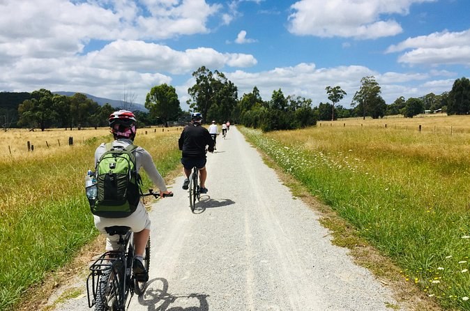 Two cyclists on a gravel rail trail between open paddocks, the ranges behind